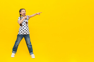 A little girl with beautiful long pigtails in jeans on a yellow isolated background shows your advertisement.