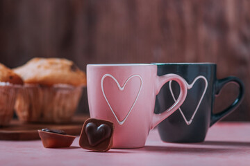 Two coffee cups with heart in pink and black colors, muffins and chocolates closeup