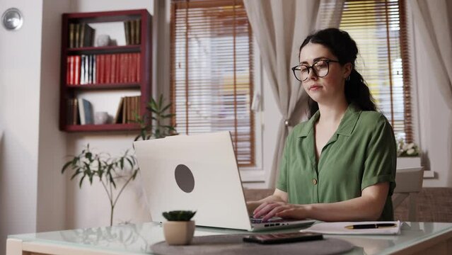 Young Caucasian pretty woman wearing eyeglasses is sitting working at laptop. Tracking right shot. Concept of freelance and remote work.