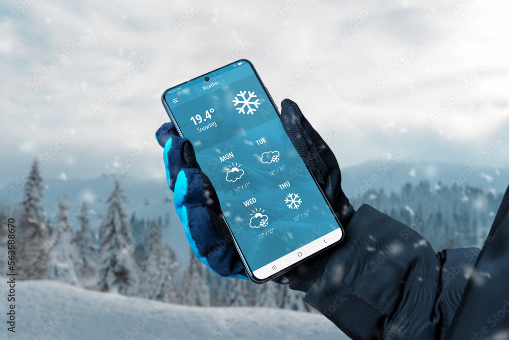 Wall mural smart phone in hand with glove with weather app on display. winter, snowing time. online service wit