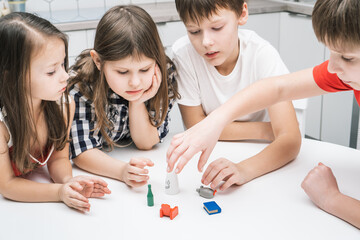 Top view of group of children playing board card game geistes blitz on white table in kitchen, stretching hand, choosing tiny figure: green bottle, red armchair, blue book, grey mouse, white ghost.