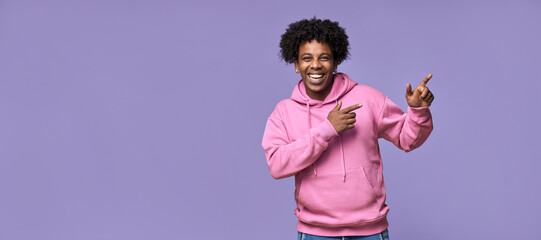 Happy African American teen guy pointing fingers aside advertising new promo offer. Smiling ethnic student model showing presenting ads standing isolated on purple background.
