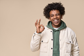 Young happy hipster African American guy student wearing white jacket isolated on beige background. Smiling cool joyful ethnic generation z teenager model laughing showing ok sign and winking. - obrazy, fototapety, plakaty