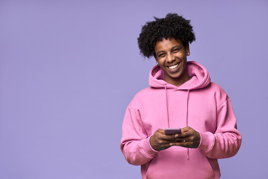 Happy cool curly African American teenage guy teen boy model wearing pink hoodie holding cell phone using mobile digital apps on cellphone texting on smartphone isolated on light purple background.