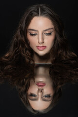 Naklejka premium Make-up and fashion concept. Brunette woman with long wavy hair sensual studio portrait. Model looking to reflection of her face in mirror. Dark studio background