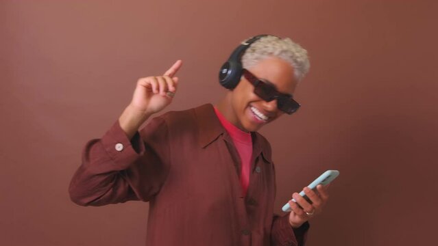 Young carefree African American woman wearing sunglasses and wireless headphones dancing enjoying cool music from new album of favorite rock band from playlist on phone posing in brown studio