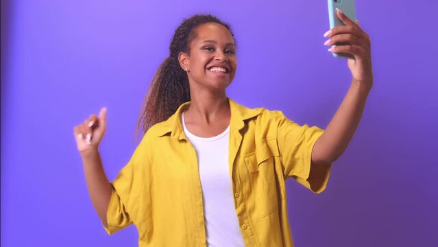 Young sociable African American woman holding phone and waving hand making video call to family or colleagues from work or recording new video for blog subscribers stands on plain purple background