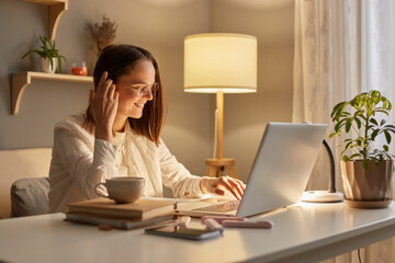 Image of smiling delighted satisfied woman wearing white shirt sitting on table and working on laptop near window, looking at pc display, expressing happiness, enjoying her work.