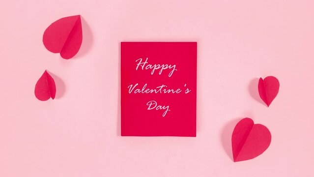 4k Near the red card with the text "Happy Valentine's Day" hearts fly in, waving their wings like butterflies. Greeting card. Pale pink background. Stop motion animation.