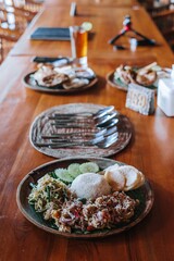 A wooden plate of Indonesian vegetable dish contains long beans, bean sprouts, cucumber, onion spice, white rice, and crackers with bokeh. Portrait or vertical.