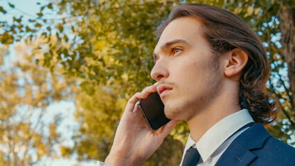Fototapeta premium Confident businessman stands outdoors with cell phone. A young businessman in a suit with smartphone. Young businessman in the park with a mobile phone.