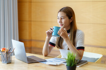Pretty Asian businesswoman sitting holding coffee at home working smiling and enjoying working with...