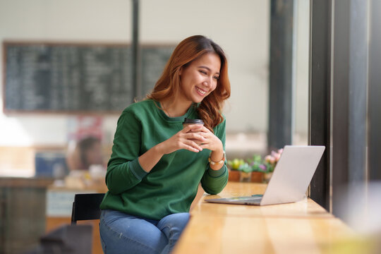 Beautiful asian businesswoman sitting and working in cafe smiling and enjoying work while having coffee and working happily with laptop computer.