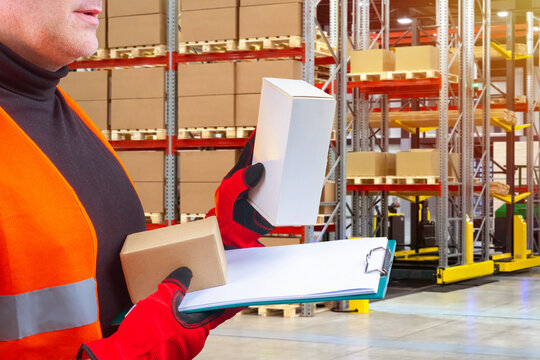 Boxes in hands of storekeeper. Man warehouse inspector with clipboard. Warehouse company supervisor holding boxes. Man in vest works in logistics warehouse. Racks with parcels on pallets behind guy