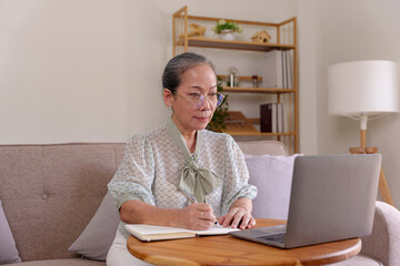 Fototapeta na wymiar Asian woman senior consultant is using laptop to chat online and take notes at work meeting at home in living room earnestly and deliberately.