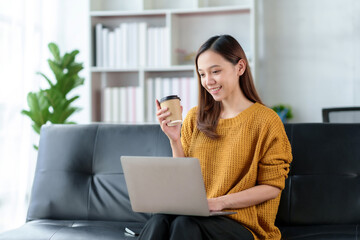 Pretty Asian businesswoman sitting holding coffee at home working smiling and enjoying working with laptop happily.