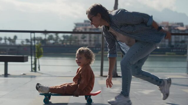 Young mother teaches ride skateboard, son of little boy, summer day. Autumnal clothes, casual clothes. Help and support parenting. Mother rides a little child on a skateboard.