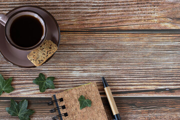 Cup of coffee, spiral notebook, pen, and green leaves on wooden table. Copy space for text, top view.