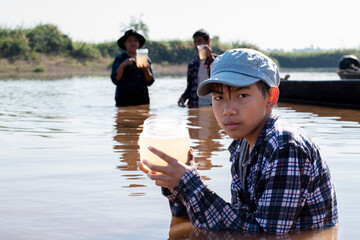 Asian boys are collecting river water samples in transparent containers to measure pH and monitor water quality and aquatic life for the report in science subject will be sent to the teacher at school