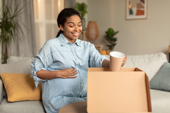 Excited Pregnant Black Woman Unpacking Cardboard Moving Box, Holding Cup And Smiling, Sitting On Couch At Home