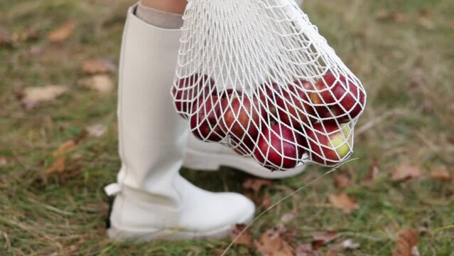 String bag with red apples. View of female legs in white boots walking through the autumn garden with eco bag in hands. Zero waste concept. No plastic life. Healthy fitness lifestyle. Recycling waste.
