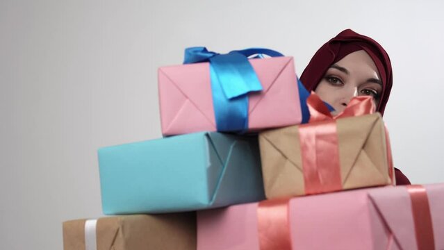 Muslim woman is carrying stack of wrapped presents in her hands which are hiding her. Isolated portrait of caucasian woman in hijab holding pile of giftboxes. Making surprise on holiday concept