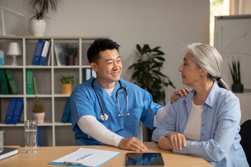 Cheerful middle aged korean man doctor supports and advises, calms old european female patient in...