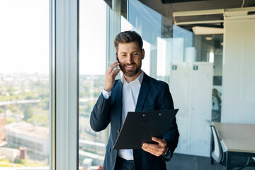 Positive middle aged businessman talking on smartphone holding file binder, standing in office near window, free space