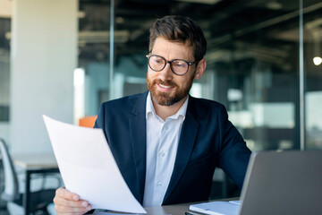 Busy middle aged businessman working with laptop and documents, sitting at desk in office, checking...