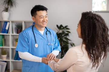 Cheerful middle aged korean doctor shaking hands, greets and supports black millennial female...
