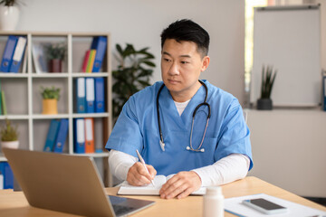 Serious middle aged korean doctor in uniform with stethoscope looks at computer, makes notes at...