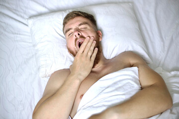 Young handsome lazy man, tired guy is yawning in bed in bedroom in the early morning, stretching hands, body. Good morning, waking up