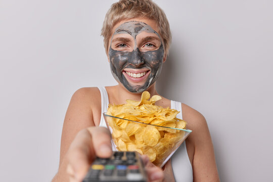 Happy woman with short hair applies clay facial mask smiles broadly switches on TV with remote control going to watch interesting film eats crisps dressed in t shirt isolated over grey background
