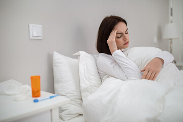 Sick woman with fever sitting in bed, touching head
