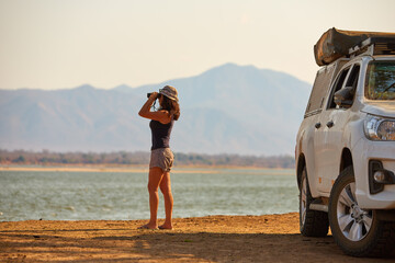 On safari in Zambia: Silhouette of a Fit Woman standing next to white safari car, observing Zambezi...