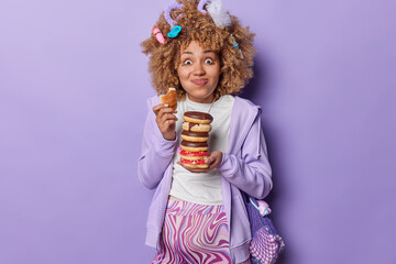 Pregnancy and overeating concept. Cheerful curly haired woman holds pile of chocolate doughnuts carries net bag poses with big belly wears sweatshirt leggings isolated over purple background
