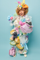 Vertical shot of emotional curly haired woman covered with plastic garbage does volunteering work clenches fists and being brave poses in full length isolated over blue wall. Environmental pollution