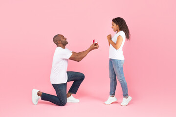 Loving black middle aged man standing on one knee and offering engagement ring to his beloved young...