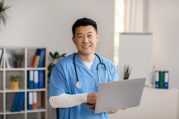 Cheerful middle aged korean doctor in uniform with stethoscope with computer in clinic office...