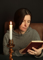A woman is reading a book, a candle is burning next to her. The woman is interested in reading, she likes to read