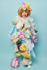 Vertical shot of emotional curly female volunteer makes costume from plastic garbage exclaims happily keeps eyes closed smiles broadly takes care of environment isolated over blue background