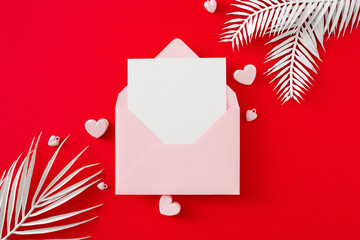 Wedding day concept. Top view photo of envelope with letter, pink hearts and white tropical leaves on red background. Lovers holiday card idea.
