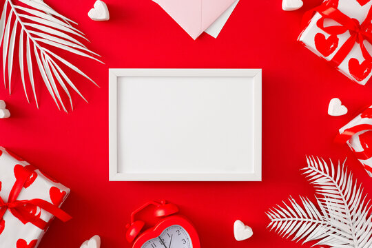 Valentines Day concept. Flat lay photo of gift boxes, white tropical leaves, envelope, alarm clock and hearts on red background with frame in the middle. Lovers holiday card idea.