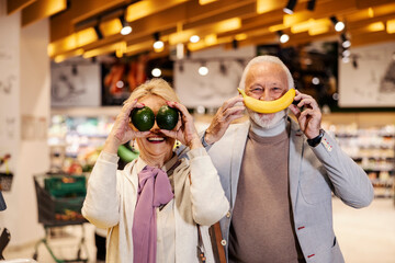 A silly senior couple is pretending fruits are their mouth and eyes while standing at the...
