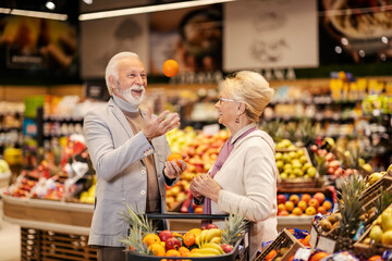 A senior man is juggling with fruits while his wife is watching him. They are purchasing fruits at the hypermarket.