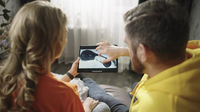 Caucasian pregnant couple looking at Uzi photo on tablet. Lifestyle of a happy man and woman expecting a child. Smiling people pregnant woman looking at baby photo. High quality 4k footage