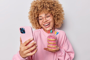 Indoor shot of curly haired woman takes selfie via smartphone celebrates birthday holds glazed doughnuts with burning candles smiles toothily wears spectacles and pullover isolated on grey background