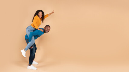 Fototapeta na wymiar Excited black man piggybacking young wife, woman pointing finger at free copy space on peach studio background, panorama