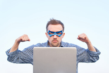 Caucasian man in nightwear shirt and superhero blue mask using laptop and showing strength gesture. Pumped fists or superpower, serious emotion concept. High quality image