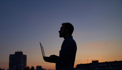 Hardly working evening. Freelance male developer or it engineer working outdoor using laptop in silhouette at sunset with urban background. Adorable dusk at background. High quality image